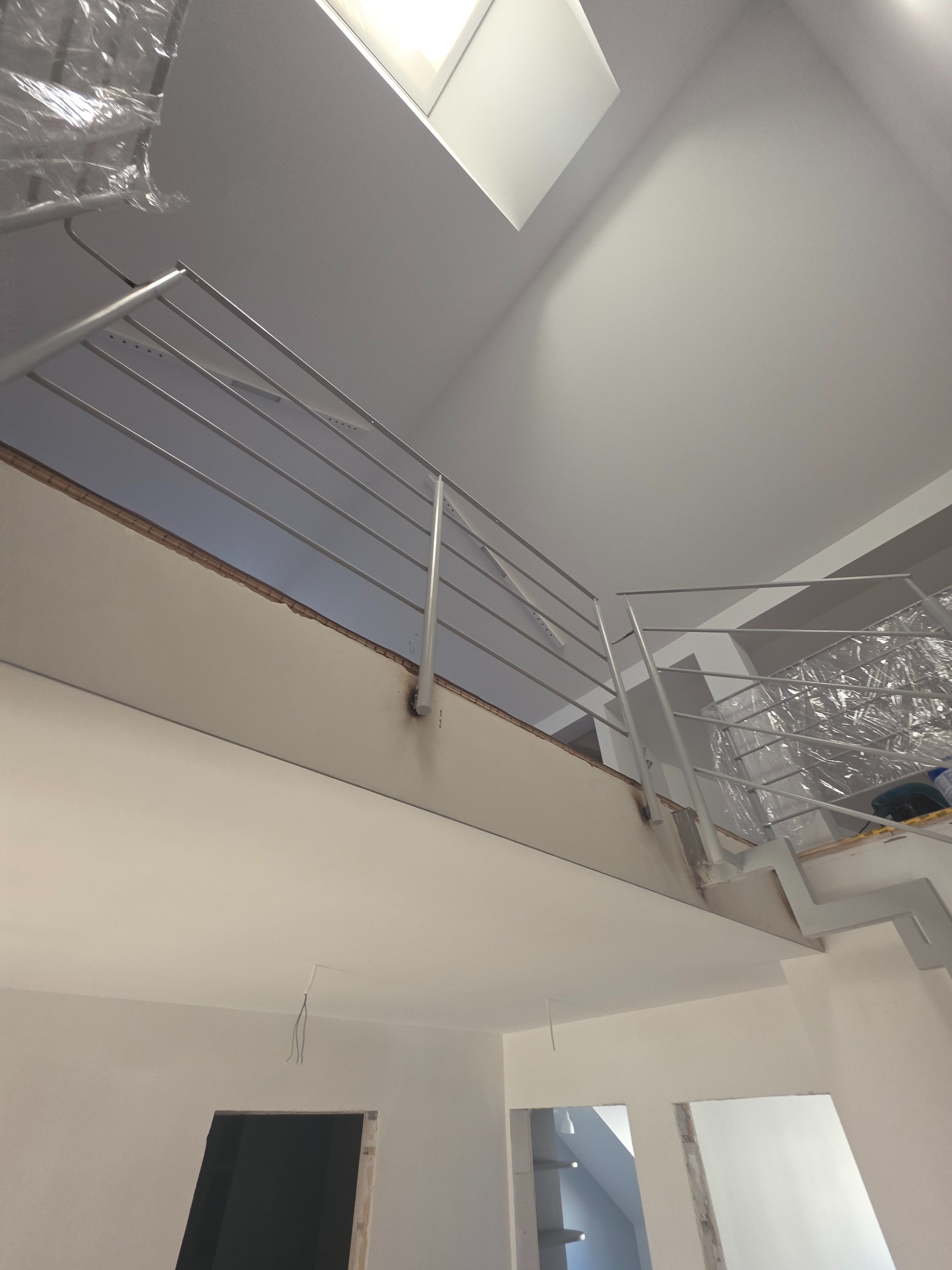 Wnętrze w trakcie remontu: białe ściany, metalowe barierki schodowe zabezpieczone folią, widok z dołu na sufit z oknem dachowym. Widoczne niedokończone otwory drzwiowe.