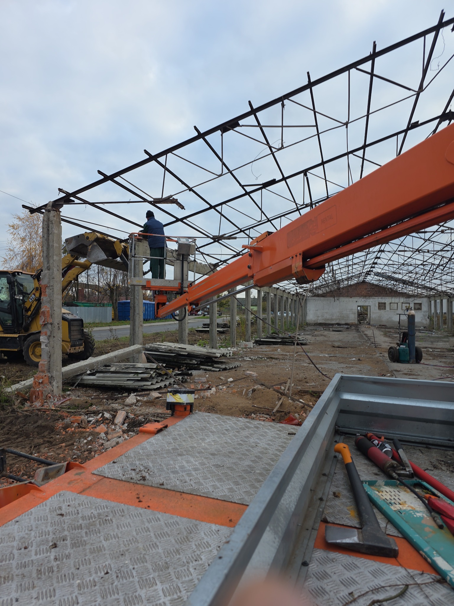 Demontaż konstrukcji hali: pracownik na podnośniku, koparka z przodu, widoczne narzędzia i fragment konstrukcji. Prace rozbiórkowe w toku, widok z góry na platformę.