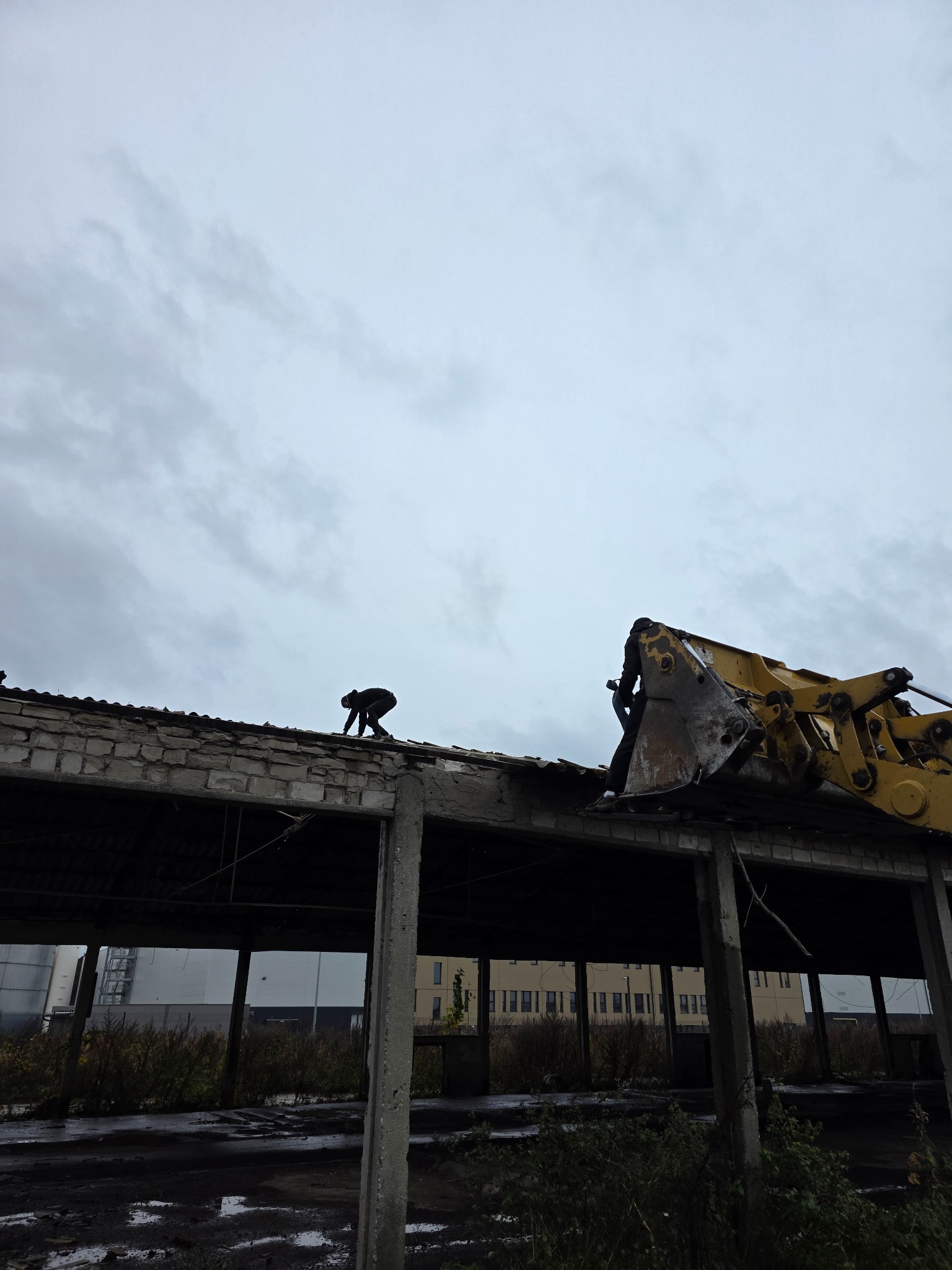 Demontaż dachu budynku z użyciem koparki. Pracownik na dachu, pochmurne niebo w tle. Widoczne elementy konstrukcyjne budynku i roślinność.