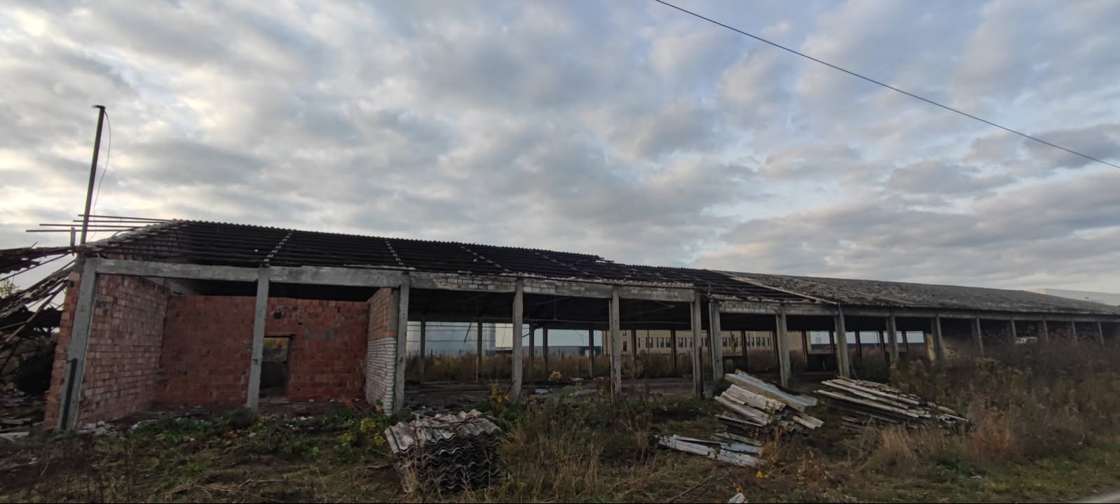 Zdjęcie zniszczonego, długiego budynku gospodarczego z częściowo zawalonym dachem i widocznymi elementami konstrukcyjnymi. W tle pochmurne niebo i zarośla.