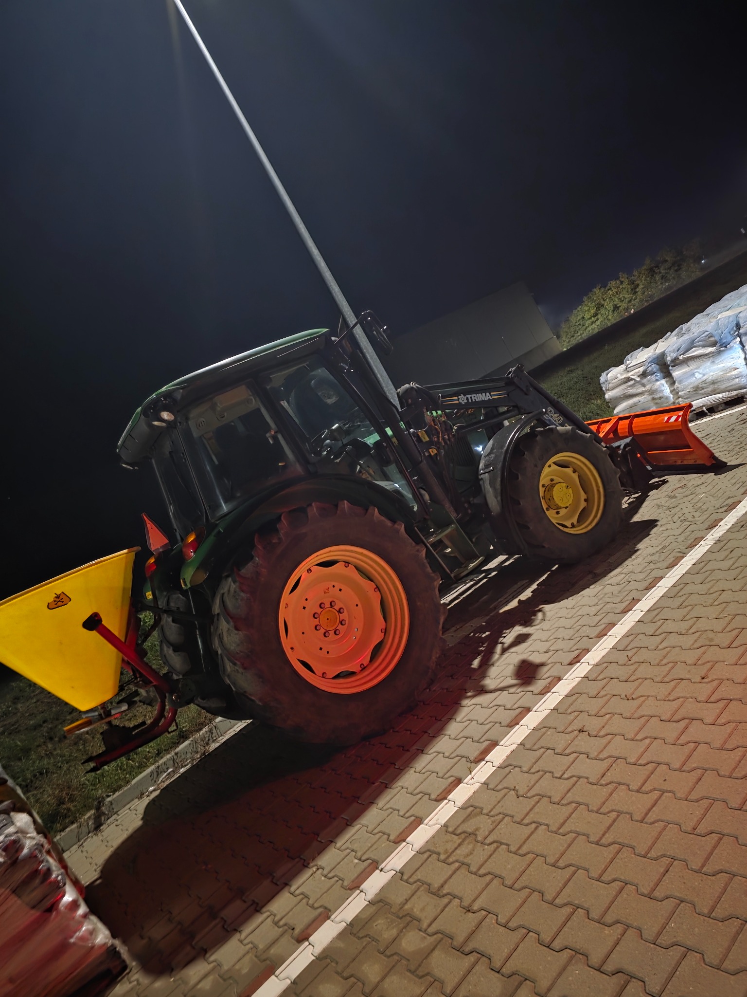 Ciężki, zielony traktor z żółtymi felgami i pomarańczowym pługiem stoi na kostce brukowej w nocy, oświetlony przez lampę. Widoczne worki w tle.