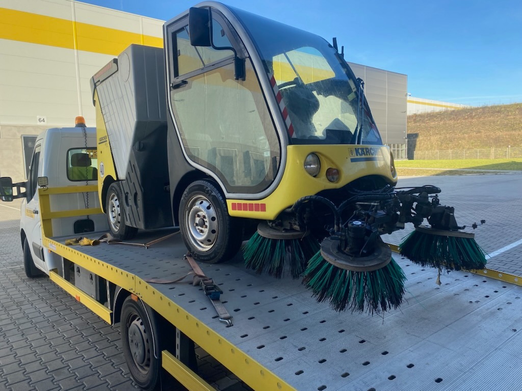 Żółto-szara zamiatarka Karcher na platformie lawety, zabezpieczona pasami. Widoczne szczotki i kabina. Tło stanowi budynek magazynowy i fragment zieleni.