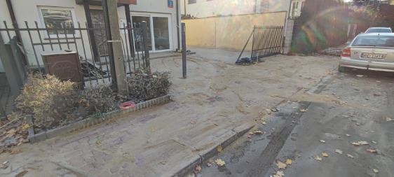 Ujęcie terenu po niwelacji, widoczne ślady opon, metalowe ogrodzenie i budynek w tle. Teren wyrównany, przygotowany pod dalsze prace.