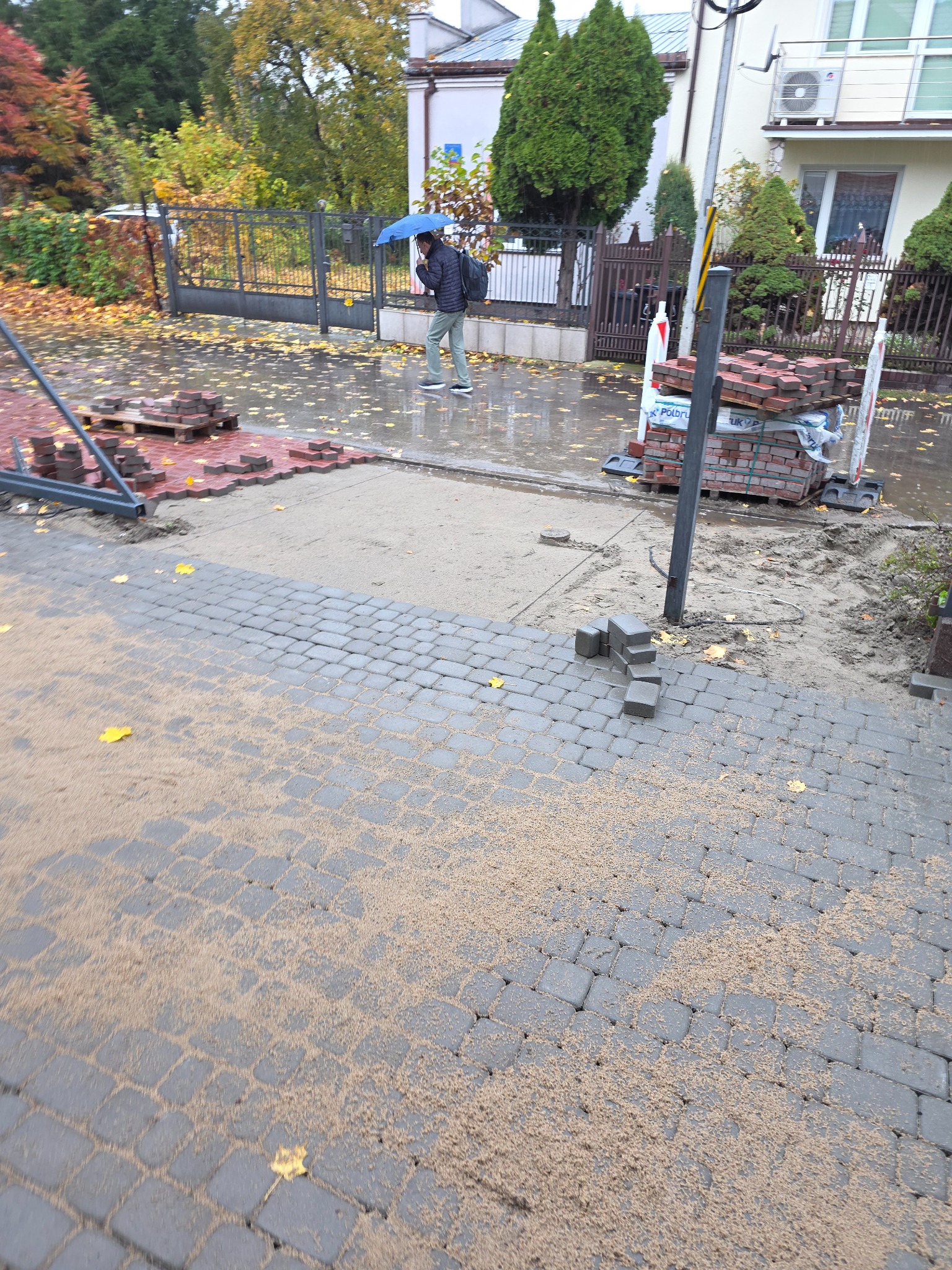 Układanie kostki brukowej na posesji w deszczowy dzień. Częściowo ułożona szara kostka, obok sterta czerwonej kostki. W tle osoba z parasolem.