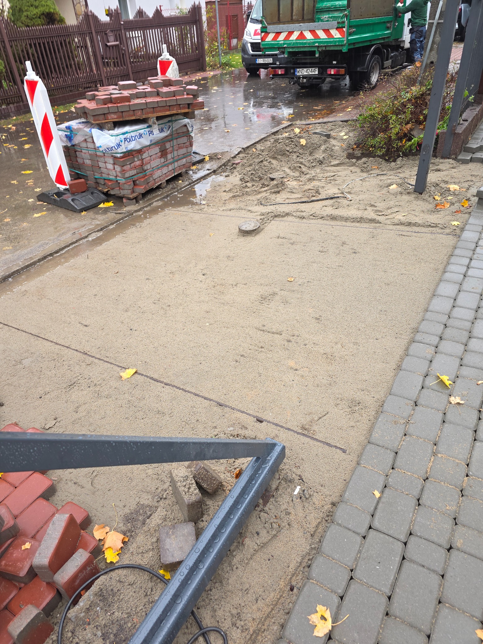 Przygotowany teren pod kostkę brukową, paleta z kostką, fragment ułożonej kostki, widoczne narzędzia i auto dostawcze w tle. Prace brukarskie w toku.