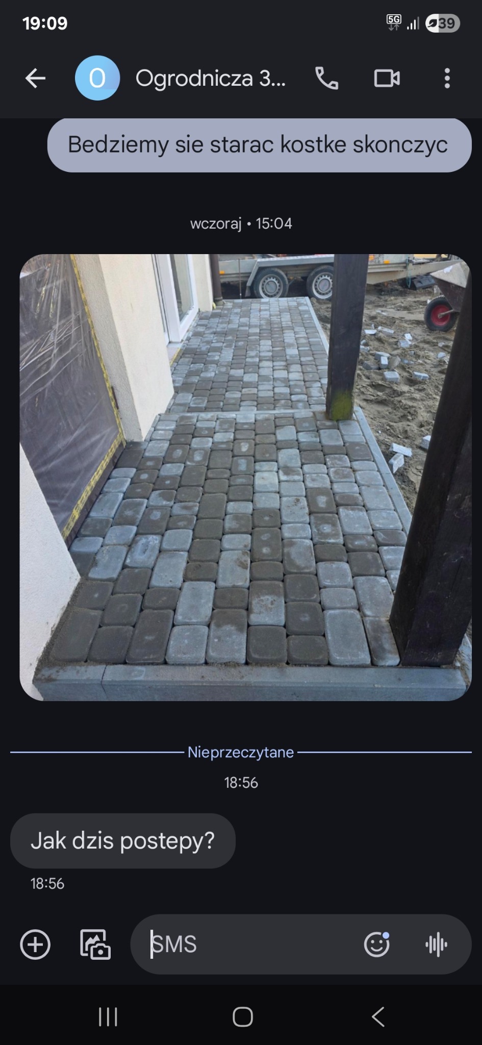 Ułożona kostka brukowa w odcieniach szarości, widok z góry, fragment budynku w tle, częściowo widoczny przyczepka i narzędzia. Zdjęcie z komunikatora.