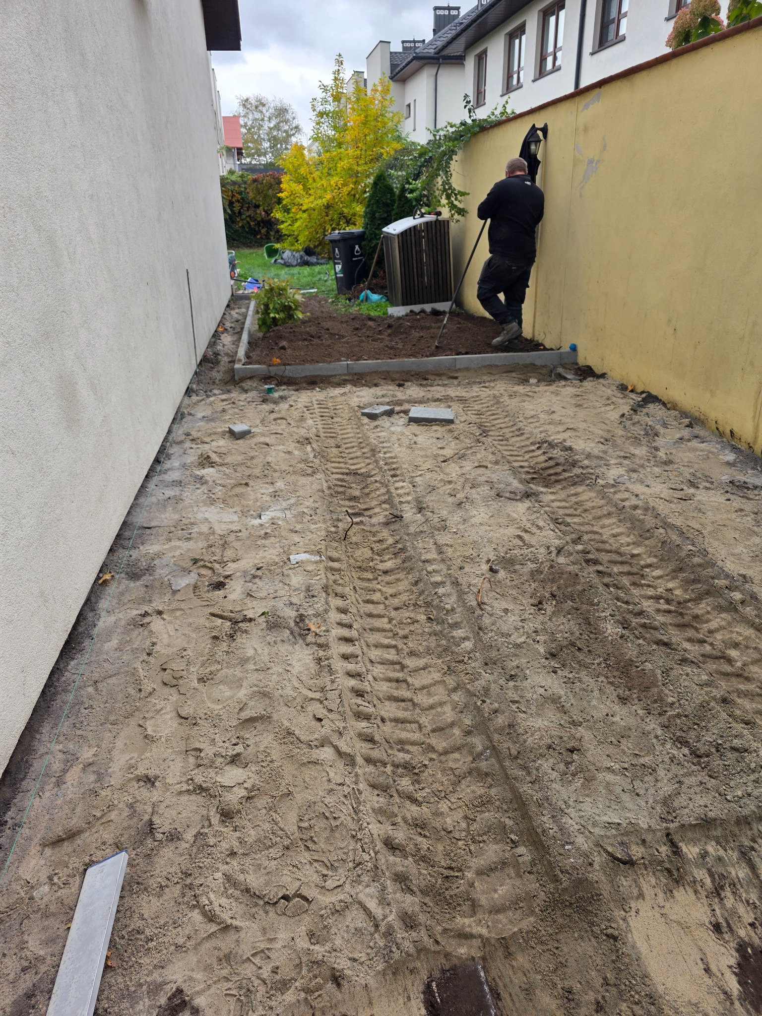 Przygotowanie terenu pod ścieżkę w ogrodzie: wyrównana ziemia z widocznymi śladami opon, obrzeża rabaty, pracownik z narzędziem przy żółtej ścianie.