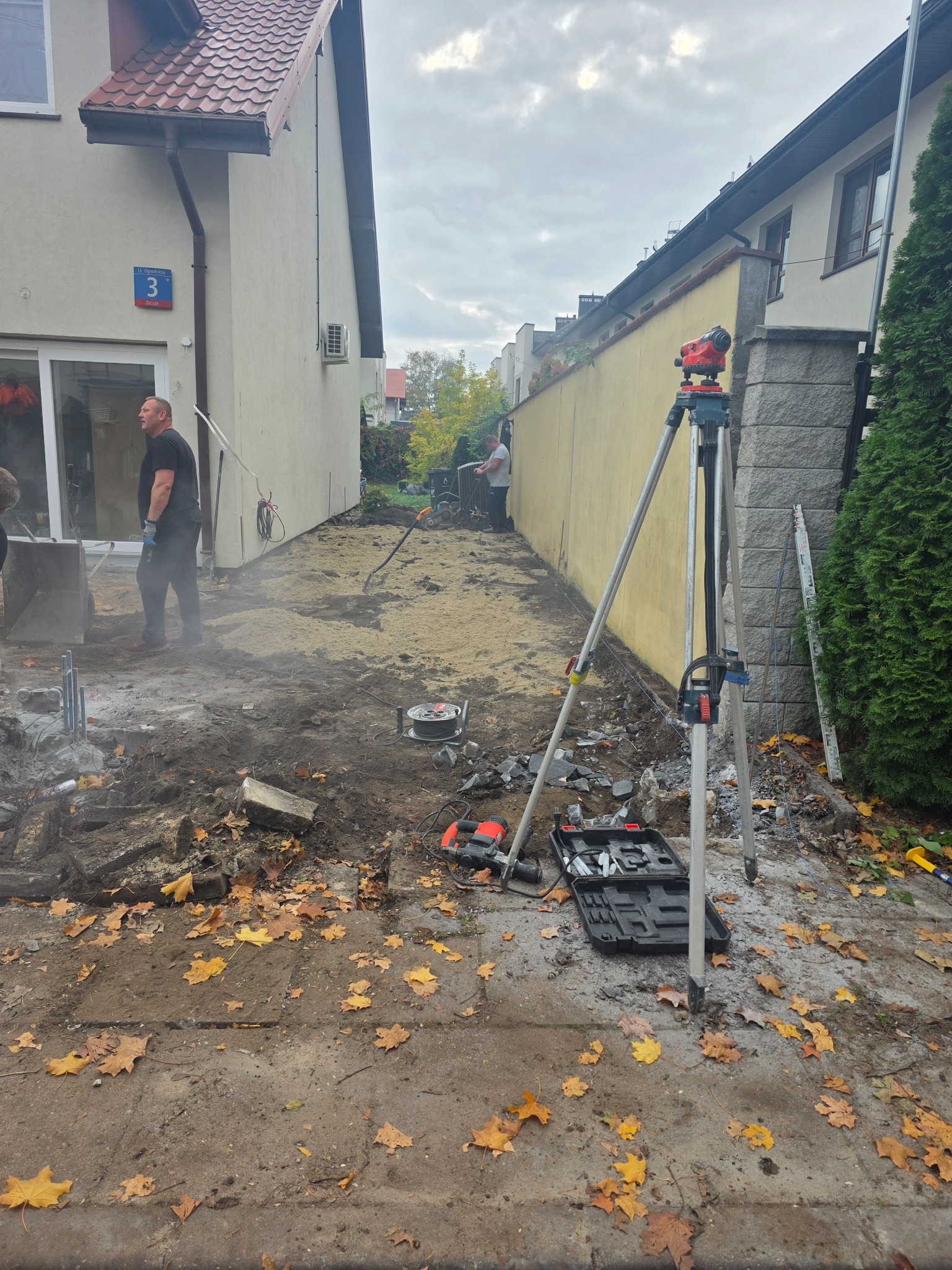 Prace ziemne w wąskim przejściu między budynkami: niwelacja terenu z użyciem niwelatora, widoczne narzędzia i robotnicy w trakcie pracy. Jesienne liście na ziemi.