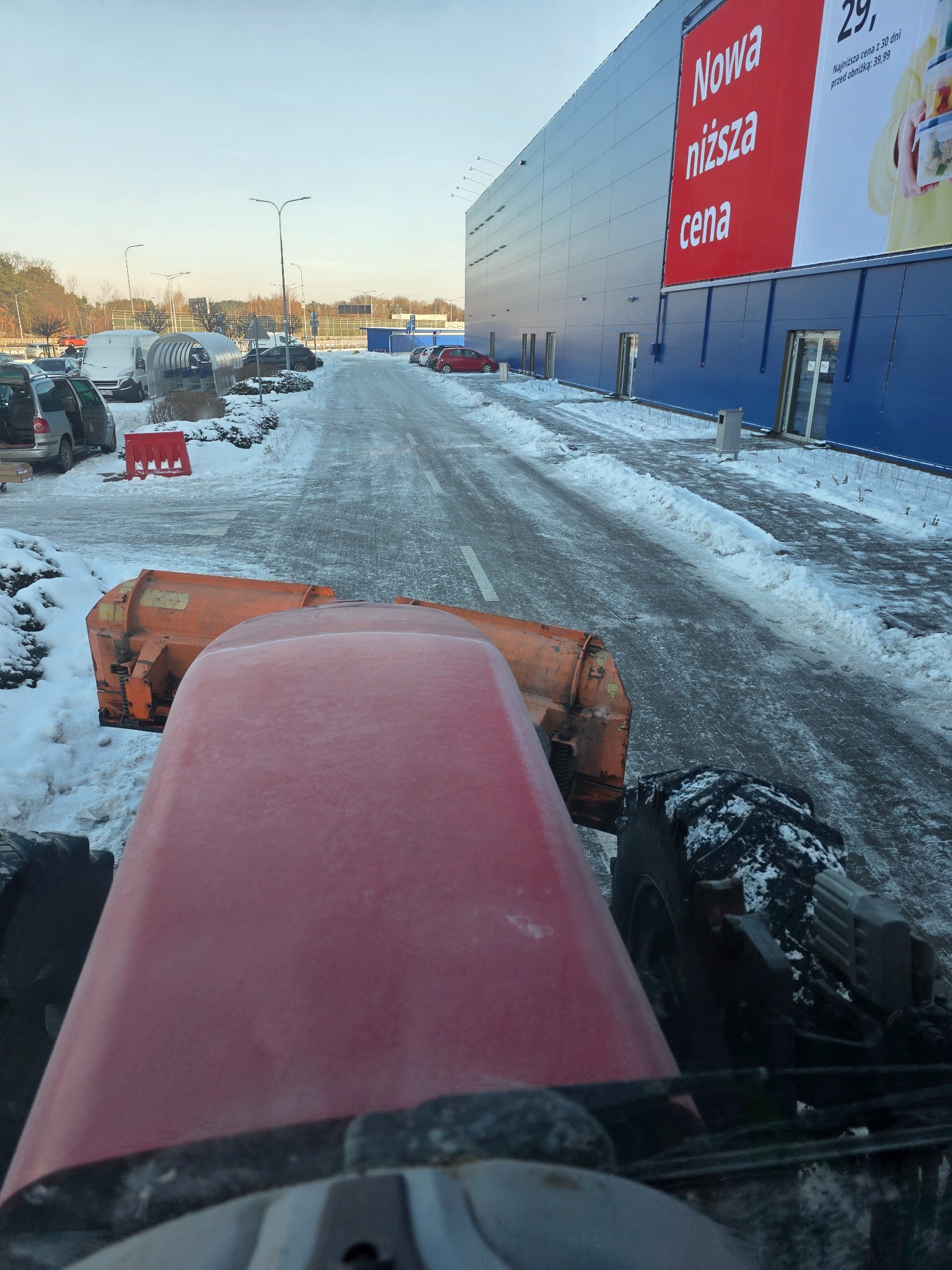 Widok z kabiny traktora z pługiem odśnieżającym, oczyszczona droga dojazdowa do budynku z reklamą, resztki śniegu po bokach, pochmurne niebo.