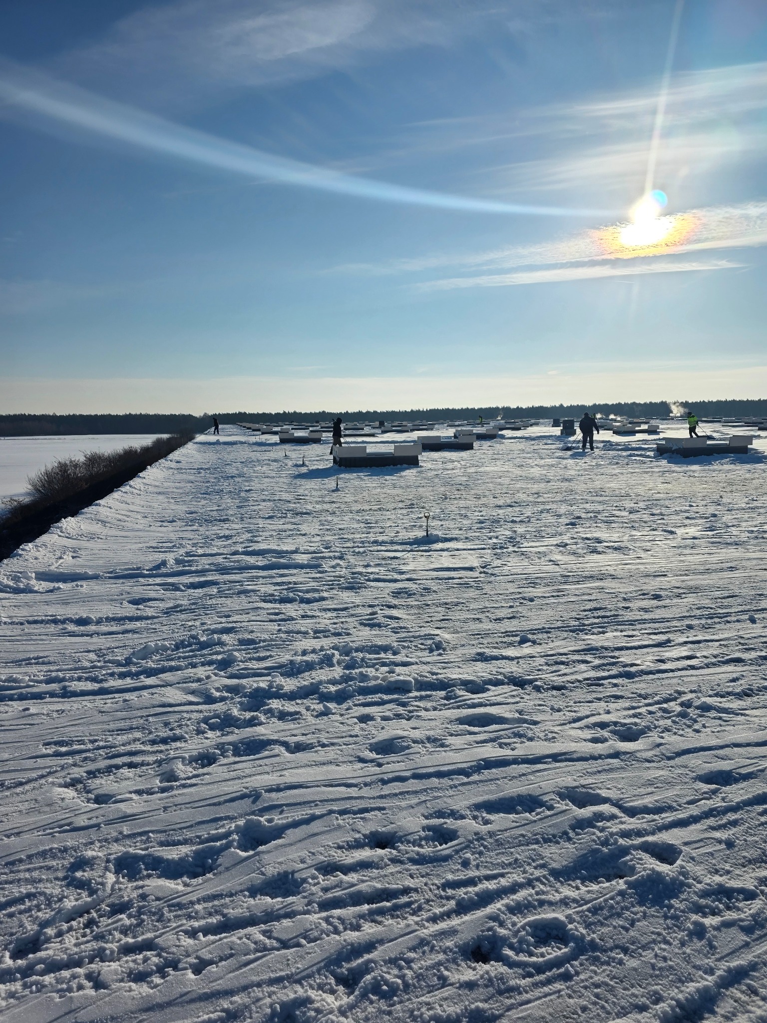 Zimowy krajobraz z grupą osób pracujących na zasypanym śniegiem terenie, prawdopodobnie przy montażu elementów konstrukcyjnych. Jasne, słoneczne niebo.