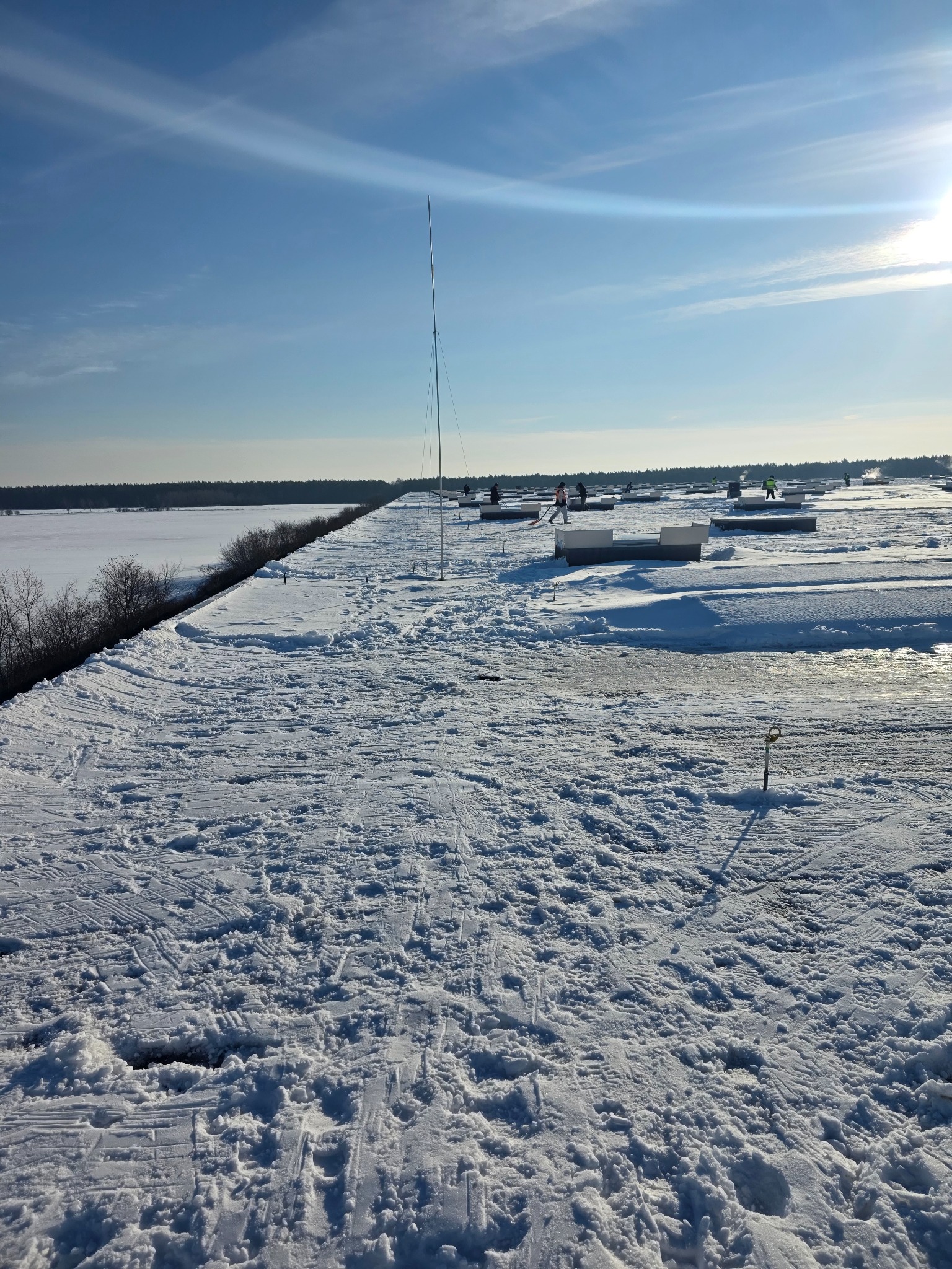 Zimowy widok z dachu budynku z anteną, gdzie pracownicy odśnieżają teren. W tle widać zamarznięte jezioro i las, a na niebie jasne słońce.
