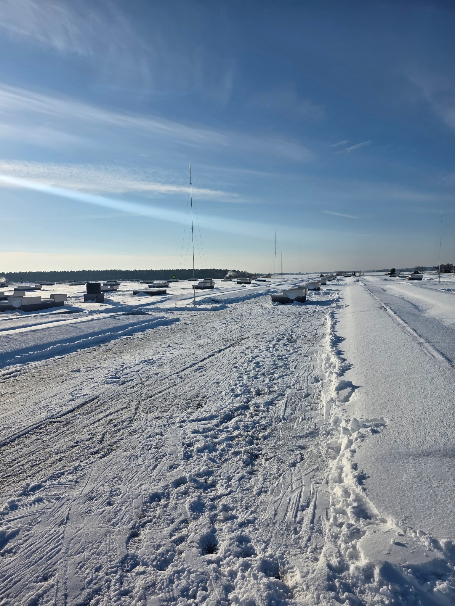 Zasypany śniegiem teren z widocznymi elementami infrastruktury i wysokimi masztami. Ślady na śniegu sugerują niedawne prace ziemne lub poruszanie się pojazdów.