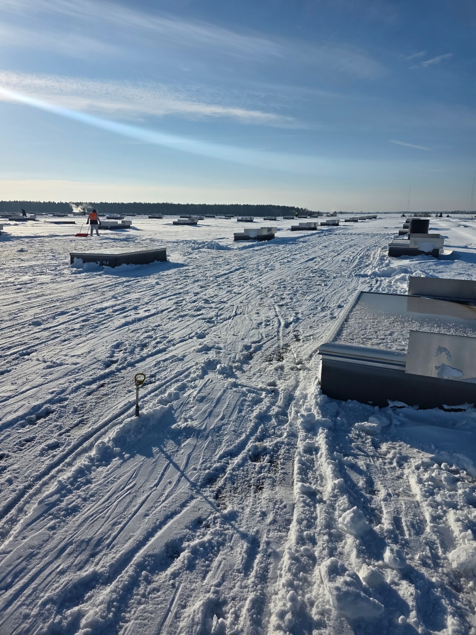 Pracownik w kamizelce odblaskowej odśnieża płaski dach pokryty śniegiem, używając czerwonej łopaty. Widoczne metalowe elementy konstrukcyjne i ślady odśnieżania.