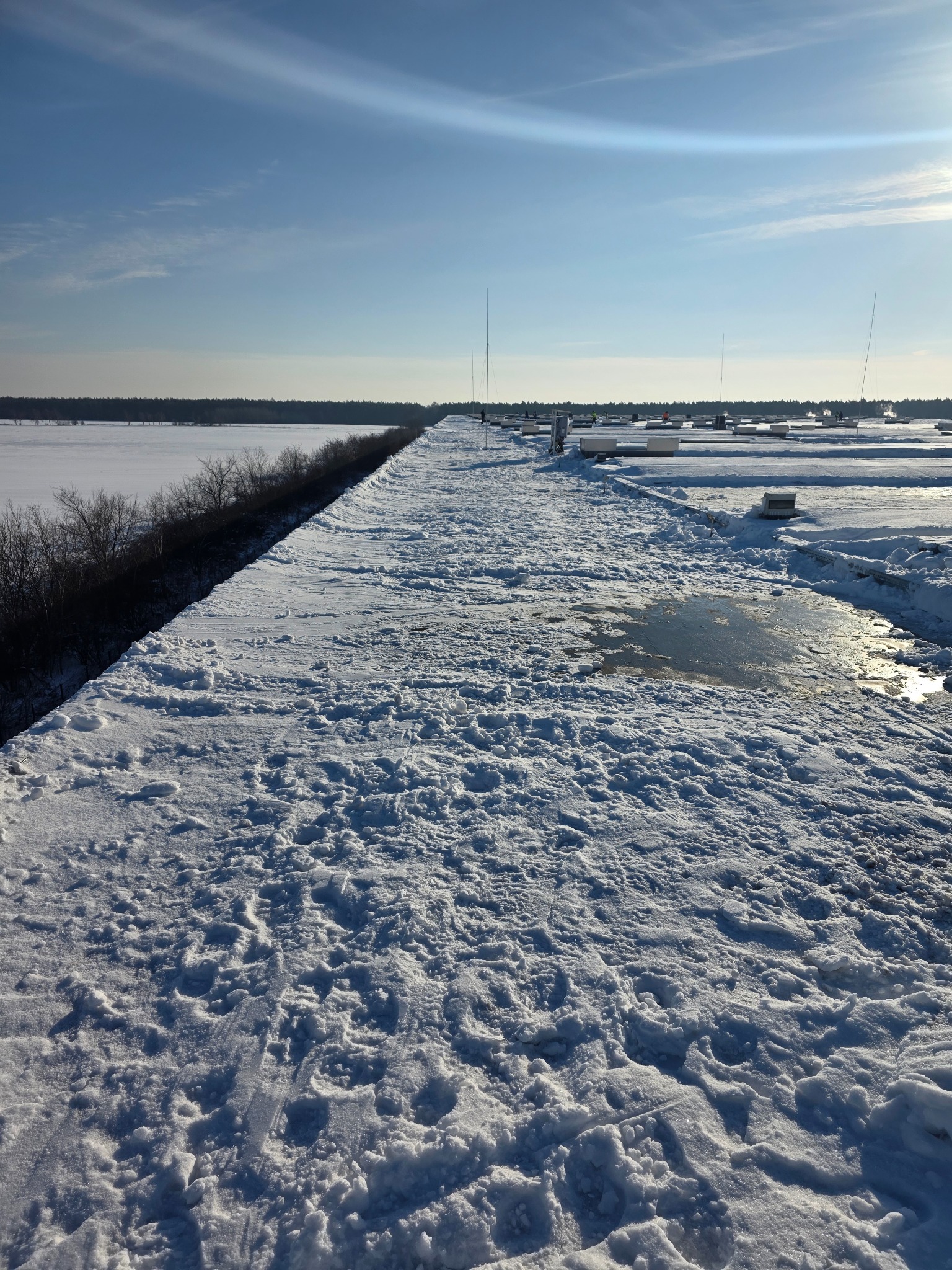 Śnieżna droga prowadząca wzdłuż brzegu jeziora, z widocznymi śladami i topniejącym śniegiem, w tle zabudowania i las pod błękitnym niebem.