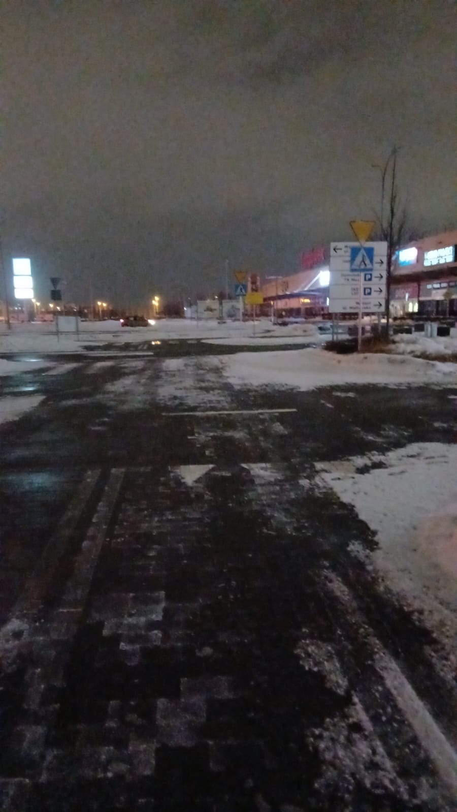 Zimowy krajobraz miejski o zmierzchu: częściowo odśnieżona droga z kostki brukowej, w tle parking z tablicami informacyjnymi i budynki handlowe z oświetleniem.