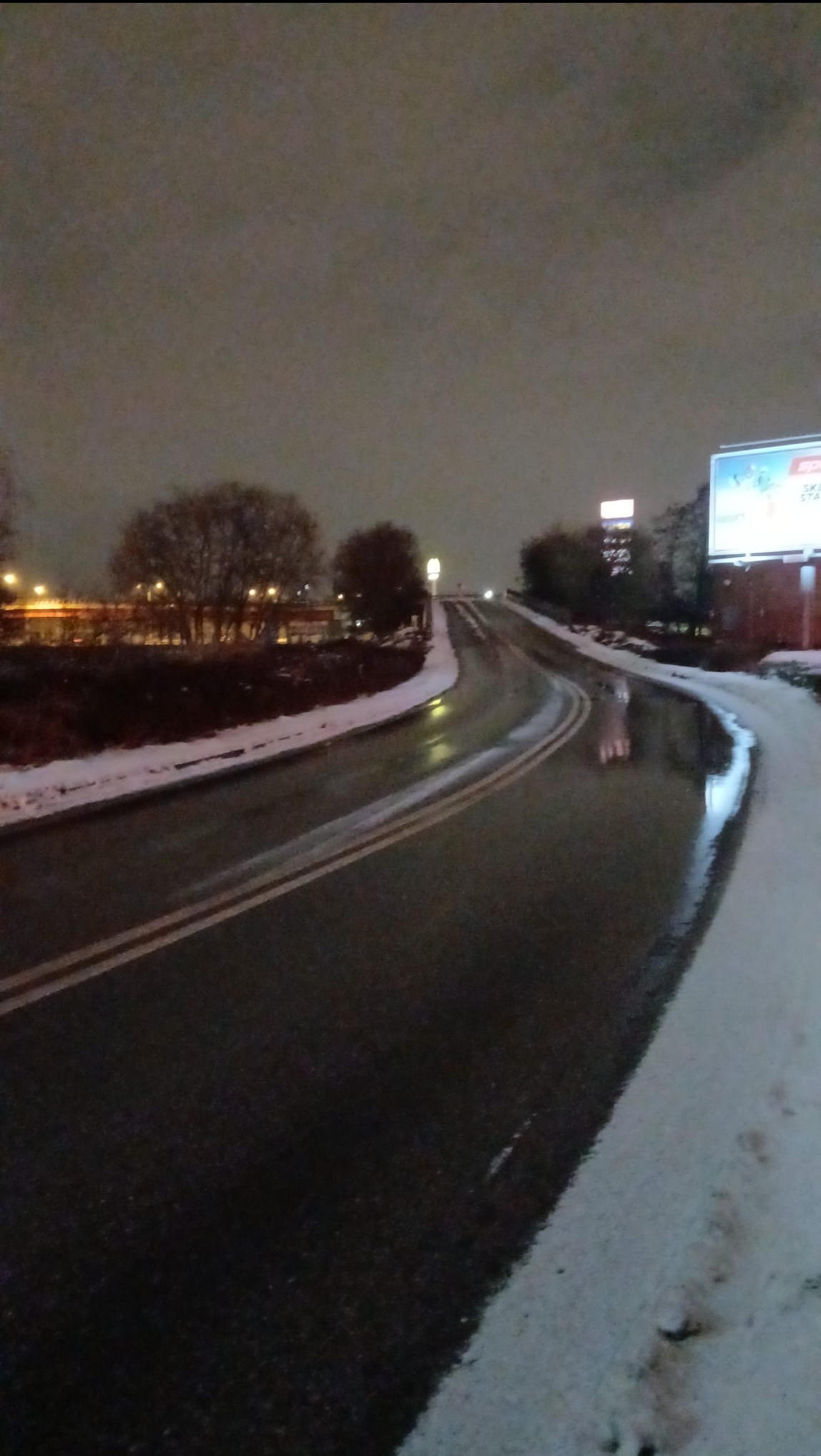 Nocna, mokra droga asfaltowa z liniami i resztkami śniegu przy krawędziach. W tle drzewa i oświetlony billboard.