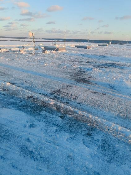 Zimowy krajobraz z betonowymi fundamentami i antenami na horyzoncie, pokryty śniegiem, pod błękitnym niebem z chmurami.