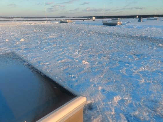 Zimowy krajobraz z zamarzniętym zbiornikiem wodnym i zabudowaniami w tle, uchwycony w chłodnym świetle poranka, z widocznym fragmentem konstrukcji w pierwszym planie.