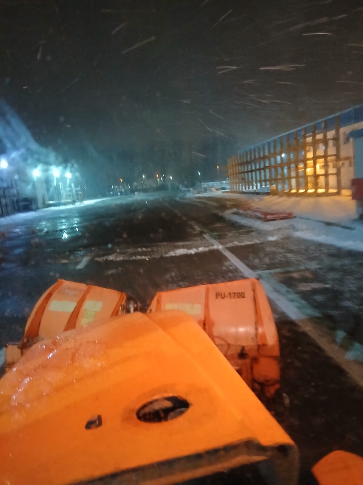 Pomarańczowy pług PU-1700 odśnieża drogę w nocy podczas intensywnych opadów śniegu. Widoczne oświetlenie uliczne i budynki w tle.