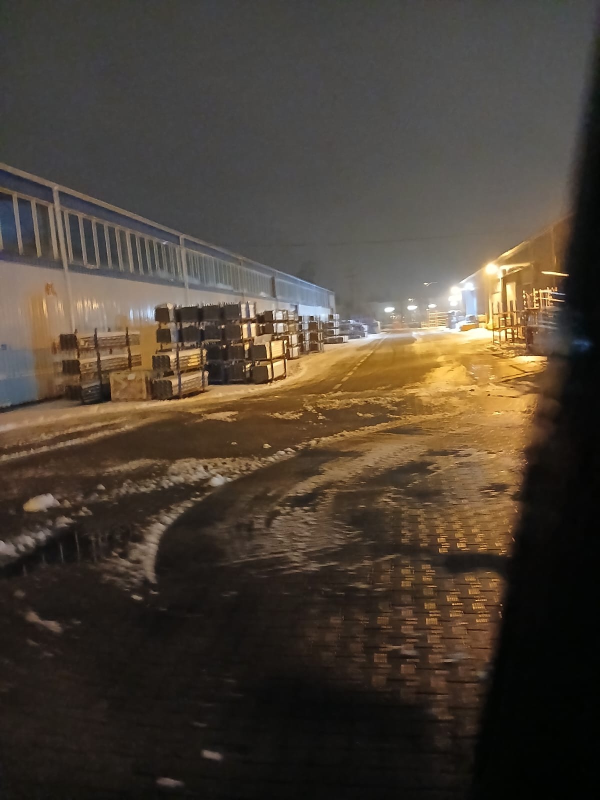 Stosy materiałów budowlanych ułożone wzdłuż ośnieżonej drogi w nocy, oświetlone przez uliczne lampy. Widoczne ślady opon na mokrej nawierzchni.