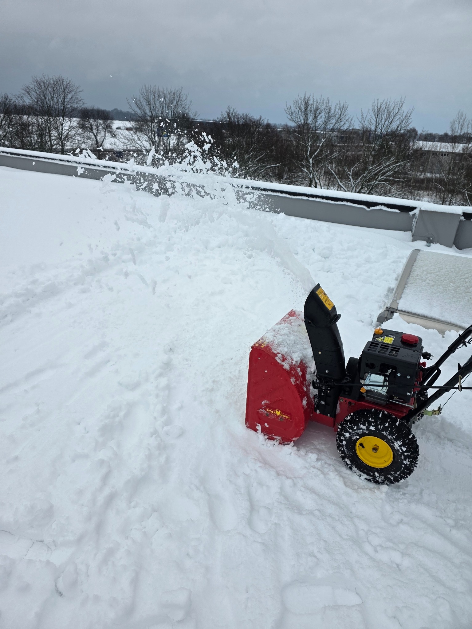 Czerwona odśnieżarka Wolf Garten w akcji, wyrzucająca śnieg na zewnątrz. Widok z góry na zasypany śniegiem dach budynku i drzewa w tle. Pochmurne niebo.