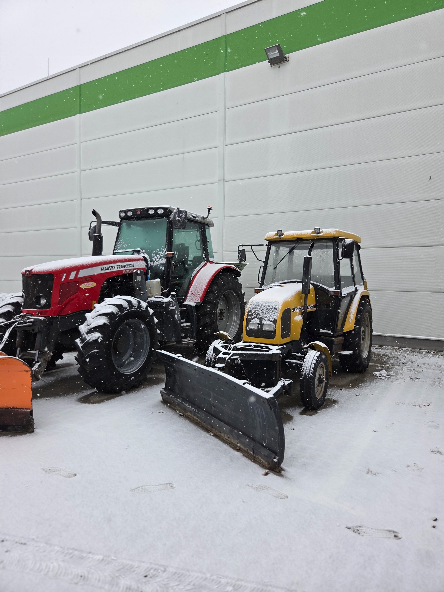Dwa traktory ze śnieżnymi pługami, czerwony Massey Ferguson i żółty Escort 450, stojące przed białą halą w zimowej scenerii. Ślady na śniegu.
