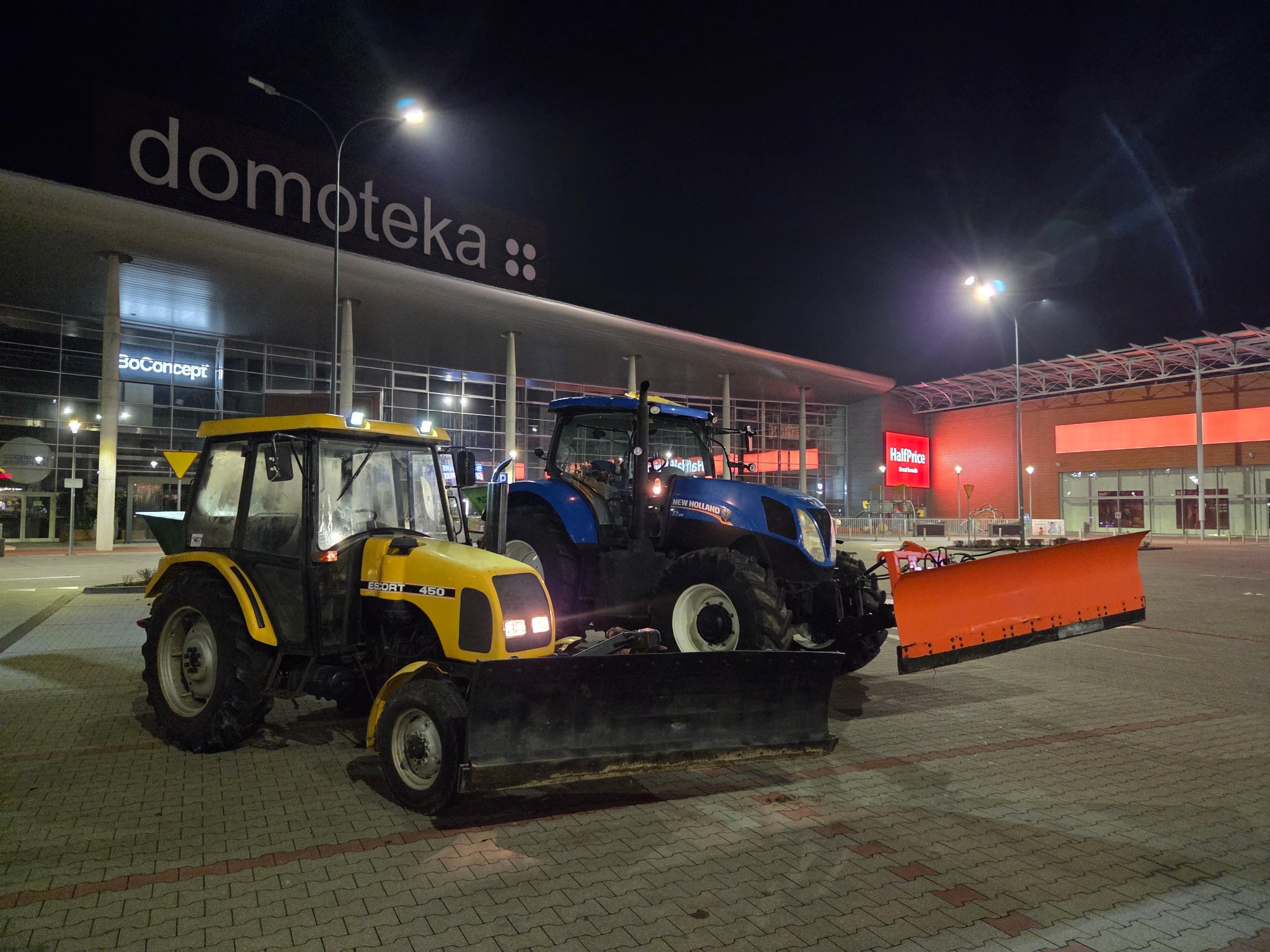 Dwa traktory z pługami do śniegu, żółty Escort 450 i niebieski New Holland, stoją na parkingu przed budynkiem Domoteki nocą, gotowe do odśnieżania.
