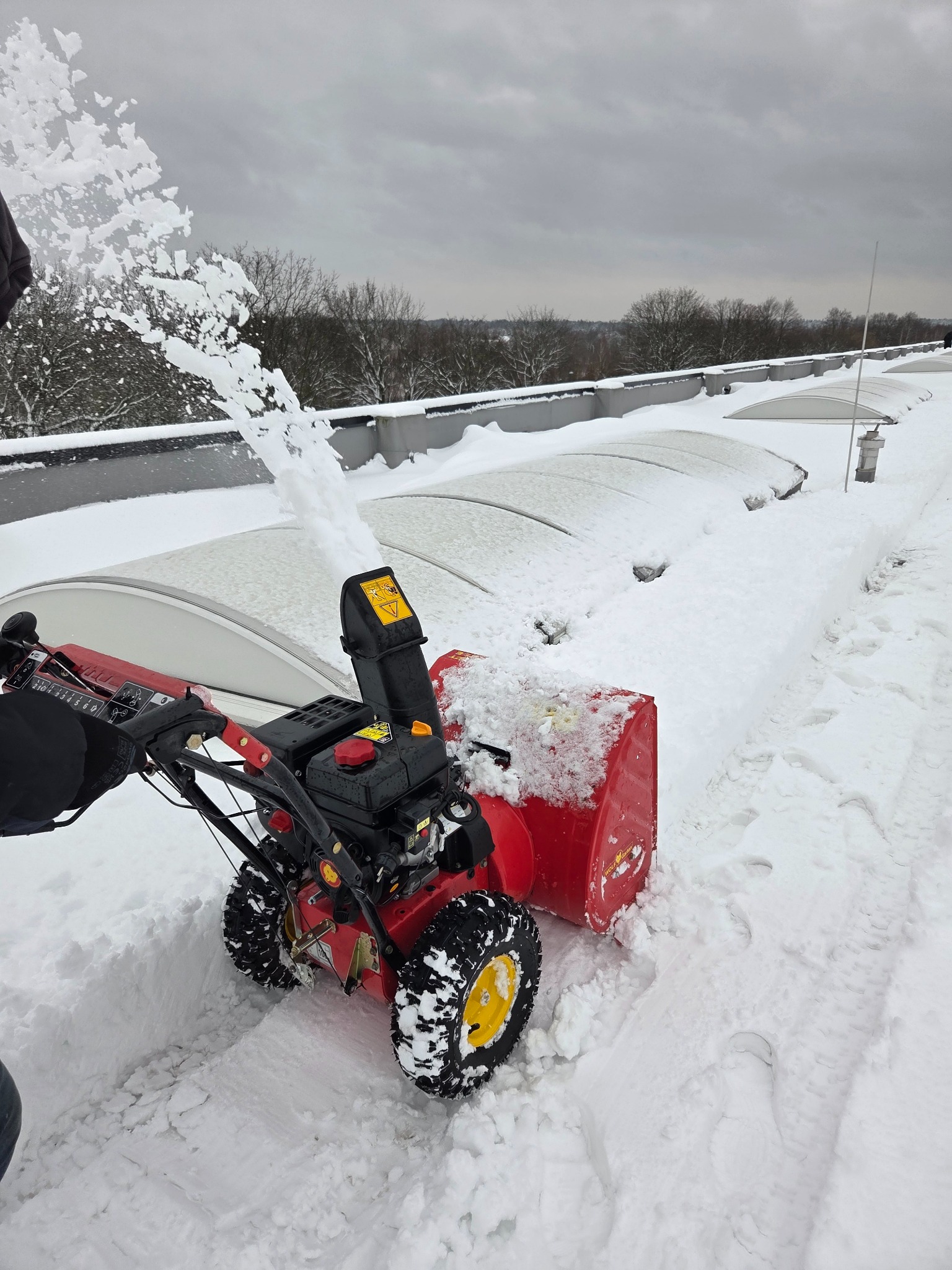 Czerwono-czarna odśnieżarka w akcji na dachu, wyrzucająca śnieg w powietrze. W tle widoczne zadaszenia i drzewa pod pochmurnym niebem.