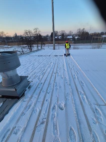Pracownik w odblaskowej kamizelce odśnieża płaski dach budynku, używając odśnieżarki. Widoczne ślady stóp w śniegu i elementy konstrukcyjne dachu.