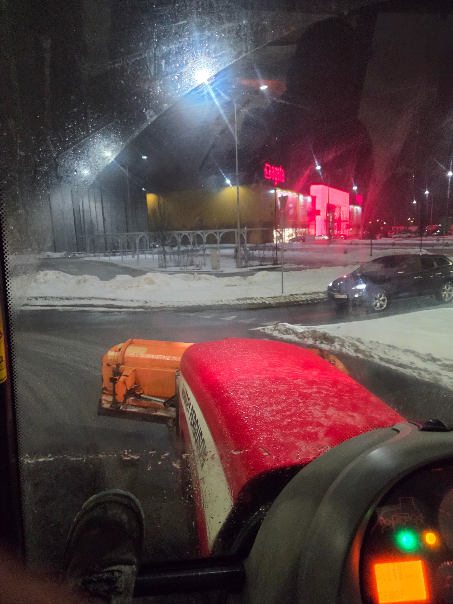 Widok z kabiny traktora Massey Ferguson odśnieżającego parking przed oświetlonym budynkiem sklepu nocą. Śnieg na drodze i chodnikach.