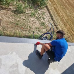 KB Budownictwo - Pracownik w czerwonych rękawicach uszczelnia szarą obróbkę blacharską dachu na tle pola i zieleni. Widok z góry, skupienie na detalu łączenia.