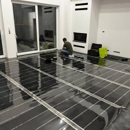 KB Budownictwo - Montaż folii grzewczej na podłodze w nowoczesnym wnętrzu. Widoczny instalator w trakcie pracy, narzędzia i rozłożone panele grzewcze.