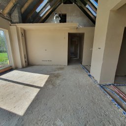 KB Budownictwo - Surowe wnętrze domu w trakcie prac wykończeniowych: widoczne instalacje podłogowe, ściany po tynkowaniu, okna dachowe. Pracownik w kombinezonie na antresoli.