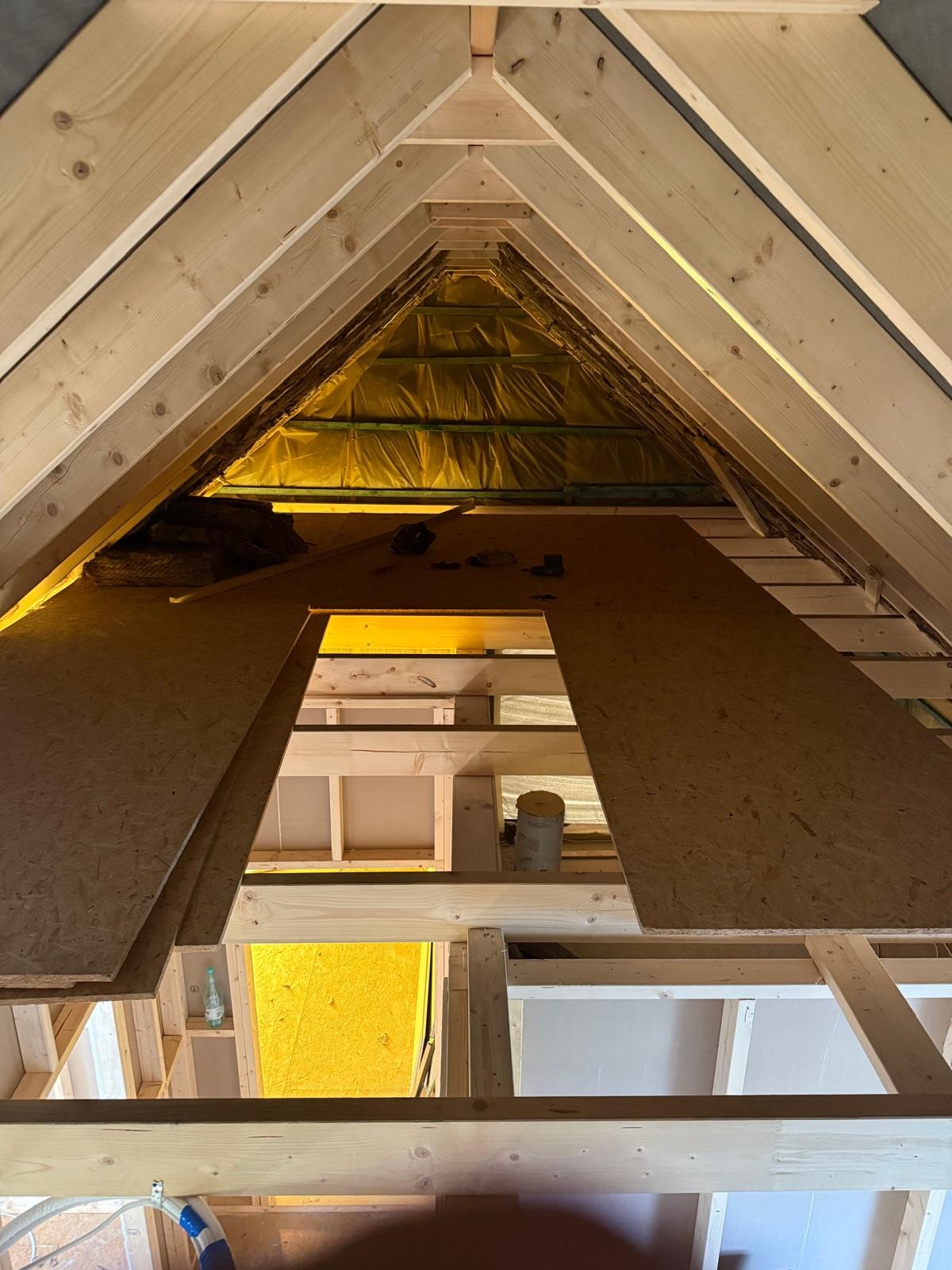 Wnętrze domu szkieletowego w trakcie budowy: widok na krokwie, izolację i płyty OSB. Konstrukcja drewniana w surowym stanie, żółta membrana w tle.