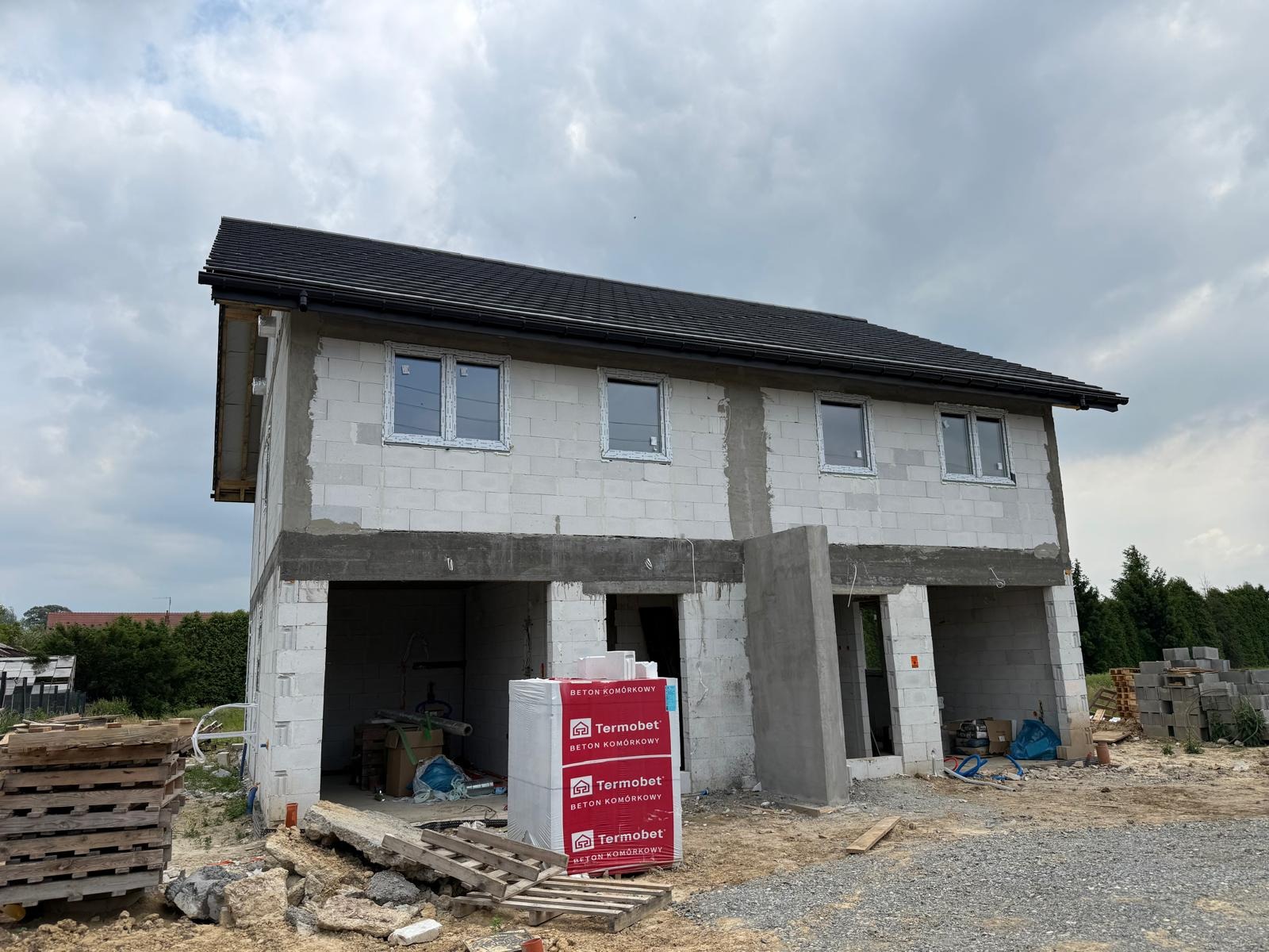 Surowy stan domu murowanego z betonu komórkowego w Michałowicach. Widoczne okna, dach i paleta bloczków Termobet. Budowa w toku, teren budowy.