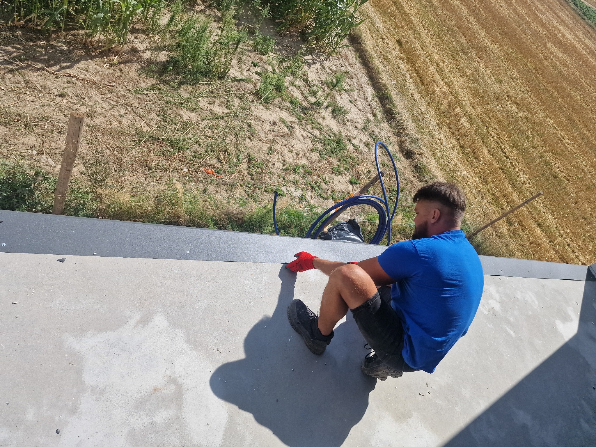 Pracownik w czerwonych rękawicach uszczelnia szarą obróbkę blacharską dachu na tle pola i zieleni. Widok z góry, skupienie na detalu łączenia.