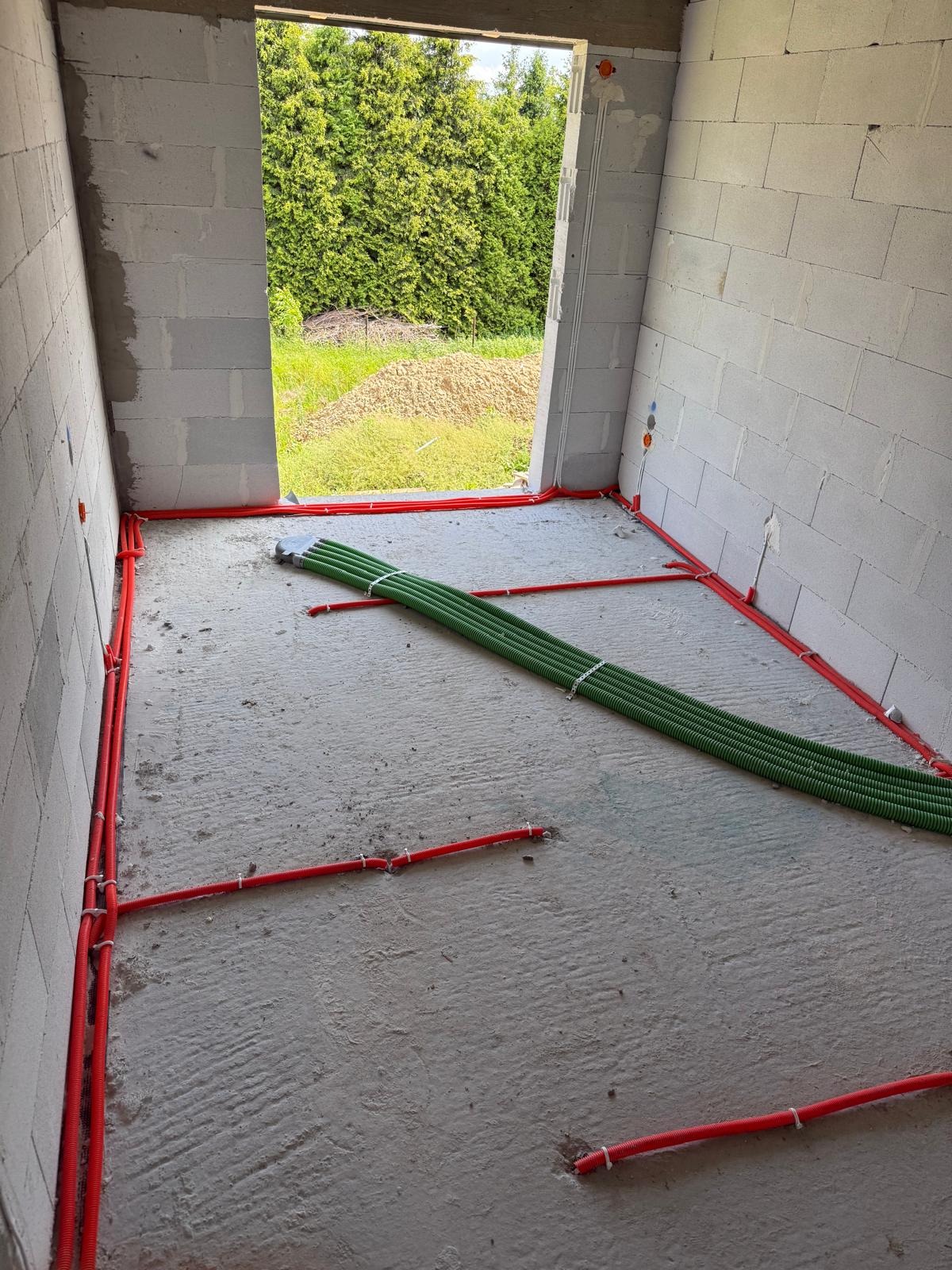 Wnętrze w trakcie budowy: instalacja elektryczna w czerwonych rurach przy ścianach i zielone rury na podłodze, widok na zewnątrz przez otwór okienny.