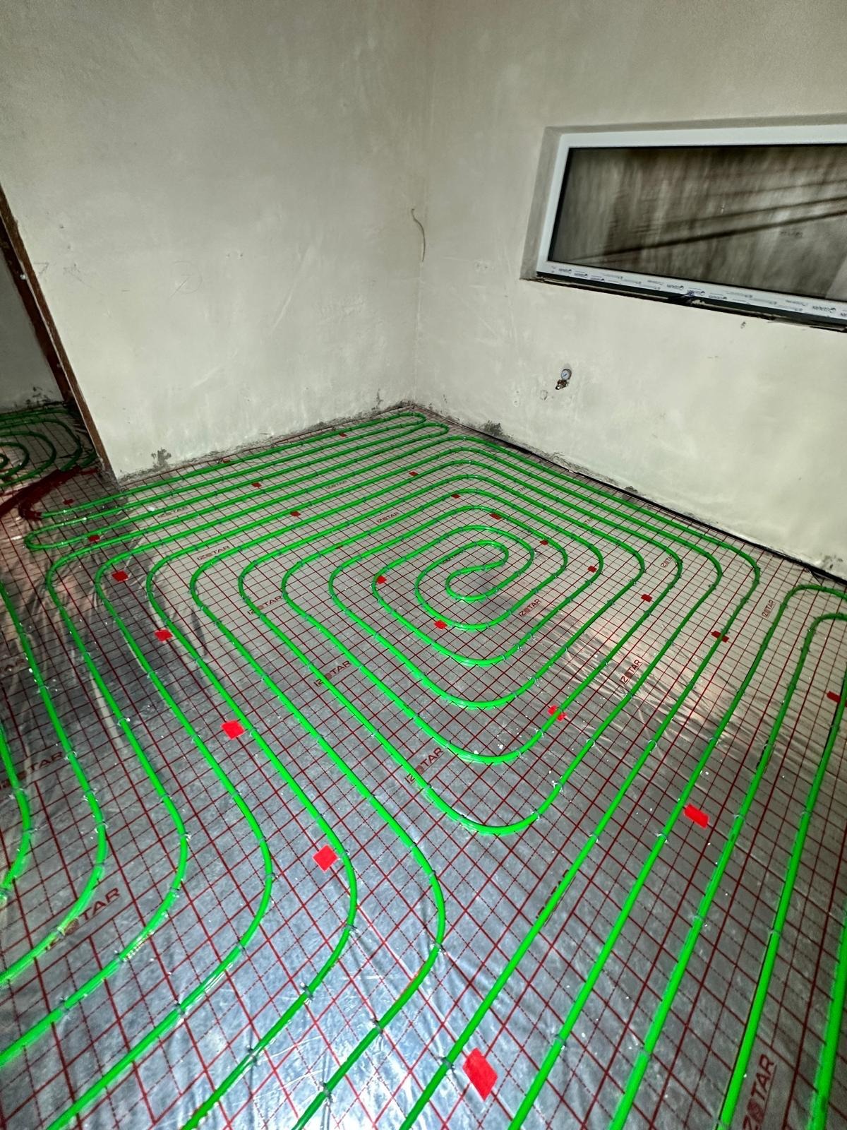 Zielone rury ogrzewania podłogowego, równomiernie rozłożone na srebrnej folii z czerwoną siatką w surowym wnętrzu z oknem i szarymi ścianami. Widoczne mocowania rur.