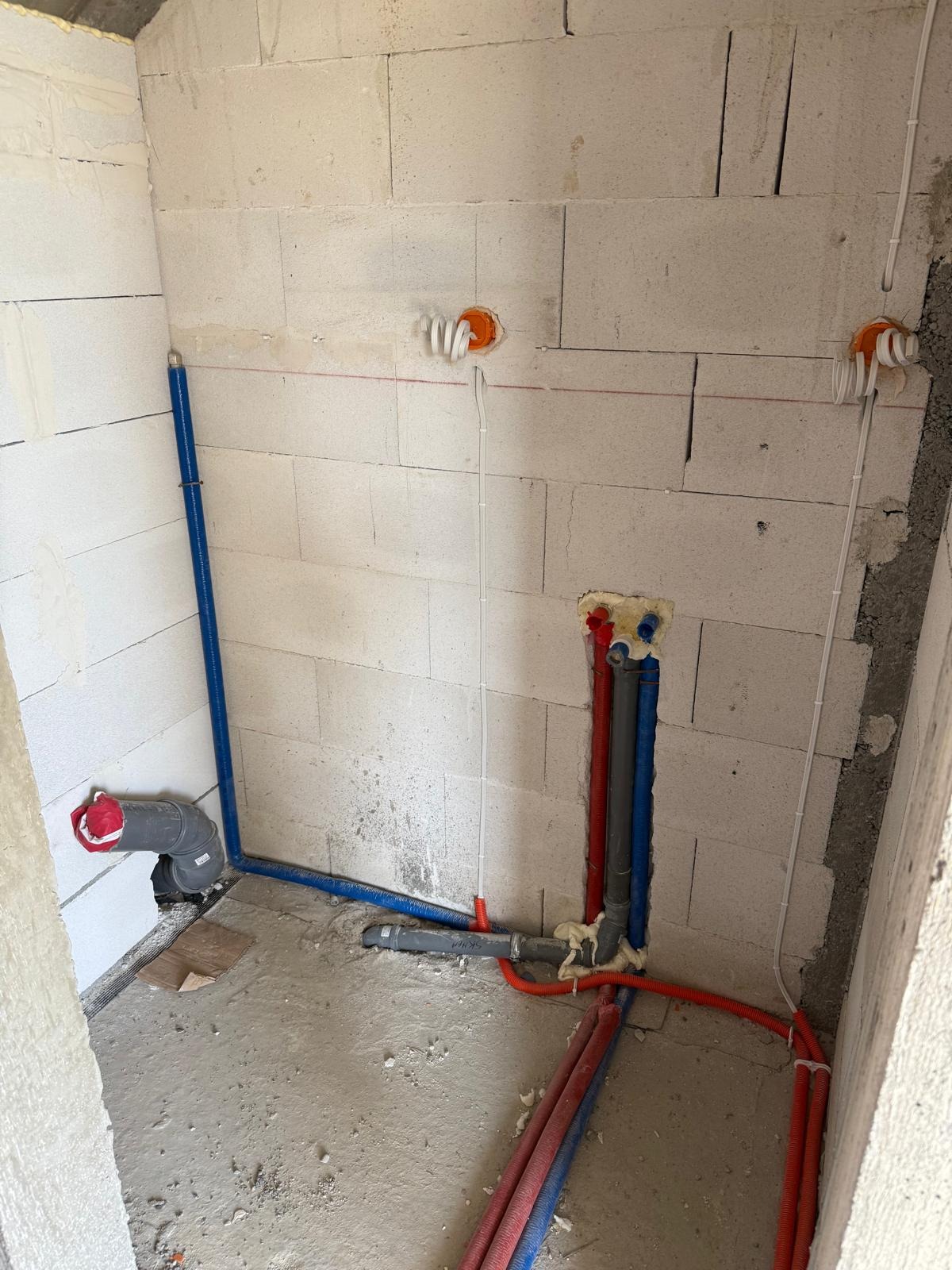 Surowy stan łazienki z instalacją wodno-kanalizacyjną i elektryczną w ścianach z betonu komórkowego. Widoczne rury i kable w kolorach niebieskim, czerwonym i pomarańczowym.