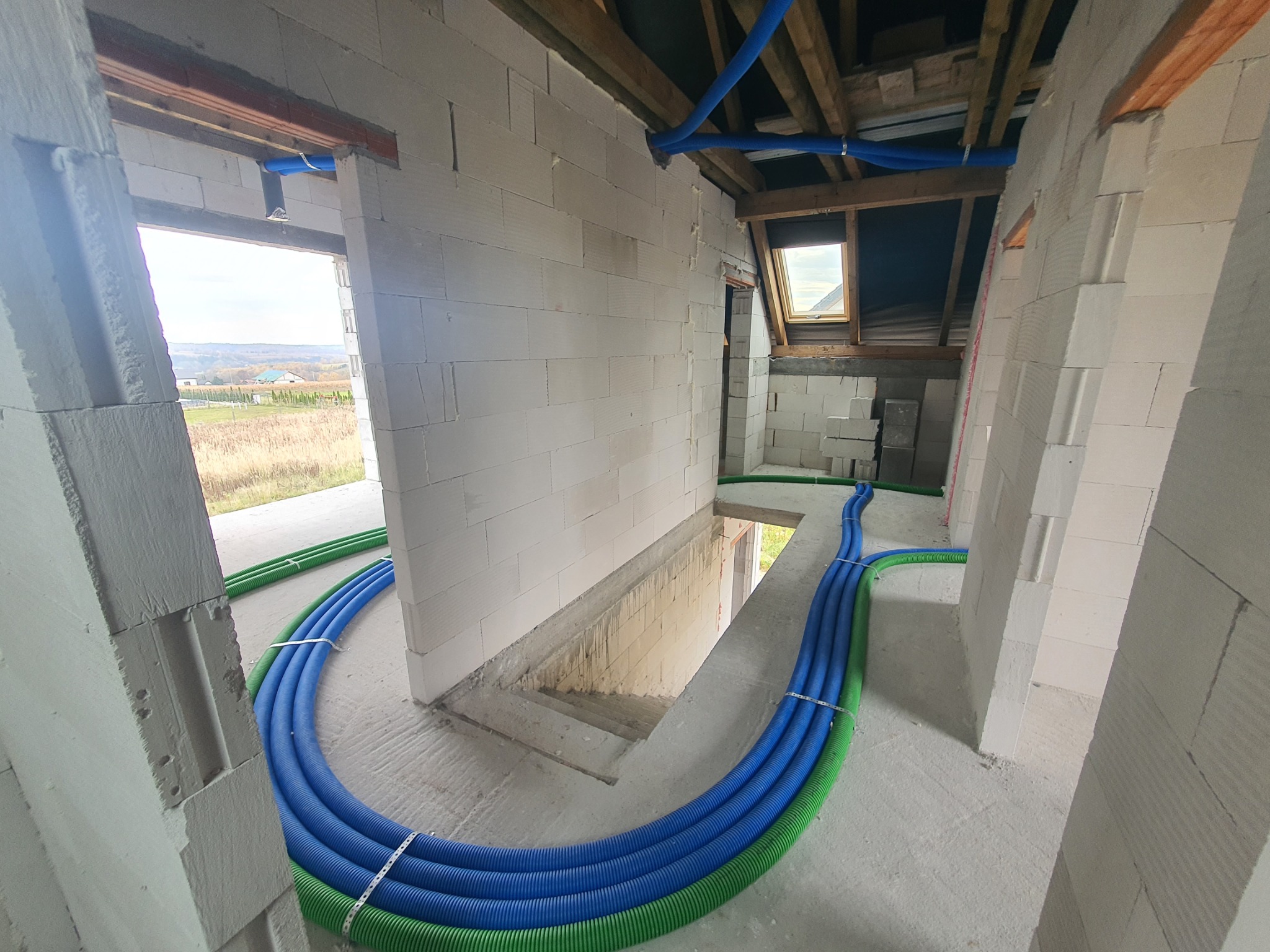 Wnętrze domu w budowie z bloczków, z widocznymi niebieskimi i zielonymi rurami instalacji w podłodze, prowadzącymi w kierunku schodów na poddasze.