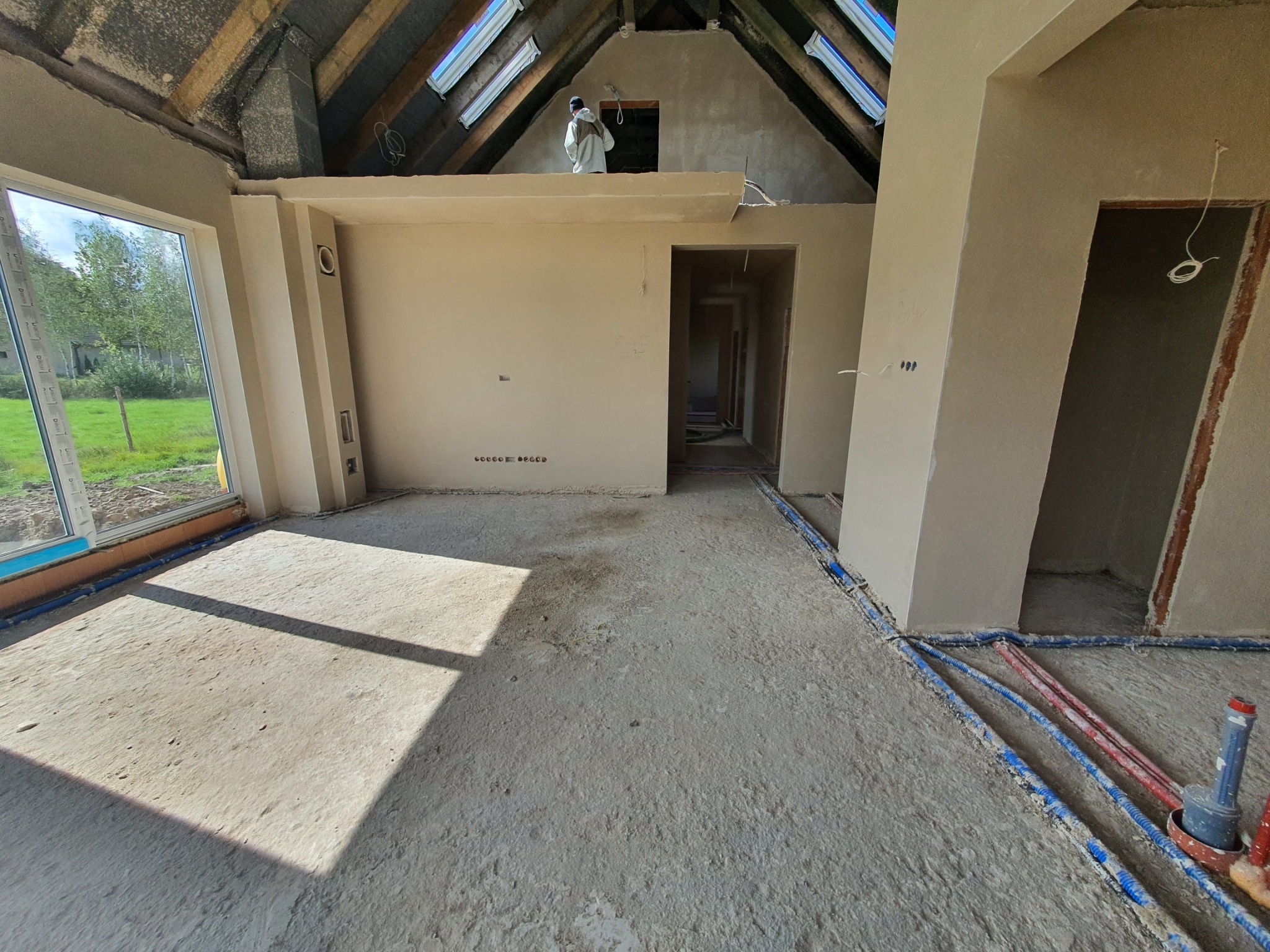 Surowe wnętrze domu w trakcie prac wykończeniowych: widoczne instalacje podłogowe, ściany po tynkowaniu, okna dachowe. Pracownik w kombinezonie na antresoli.