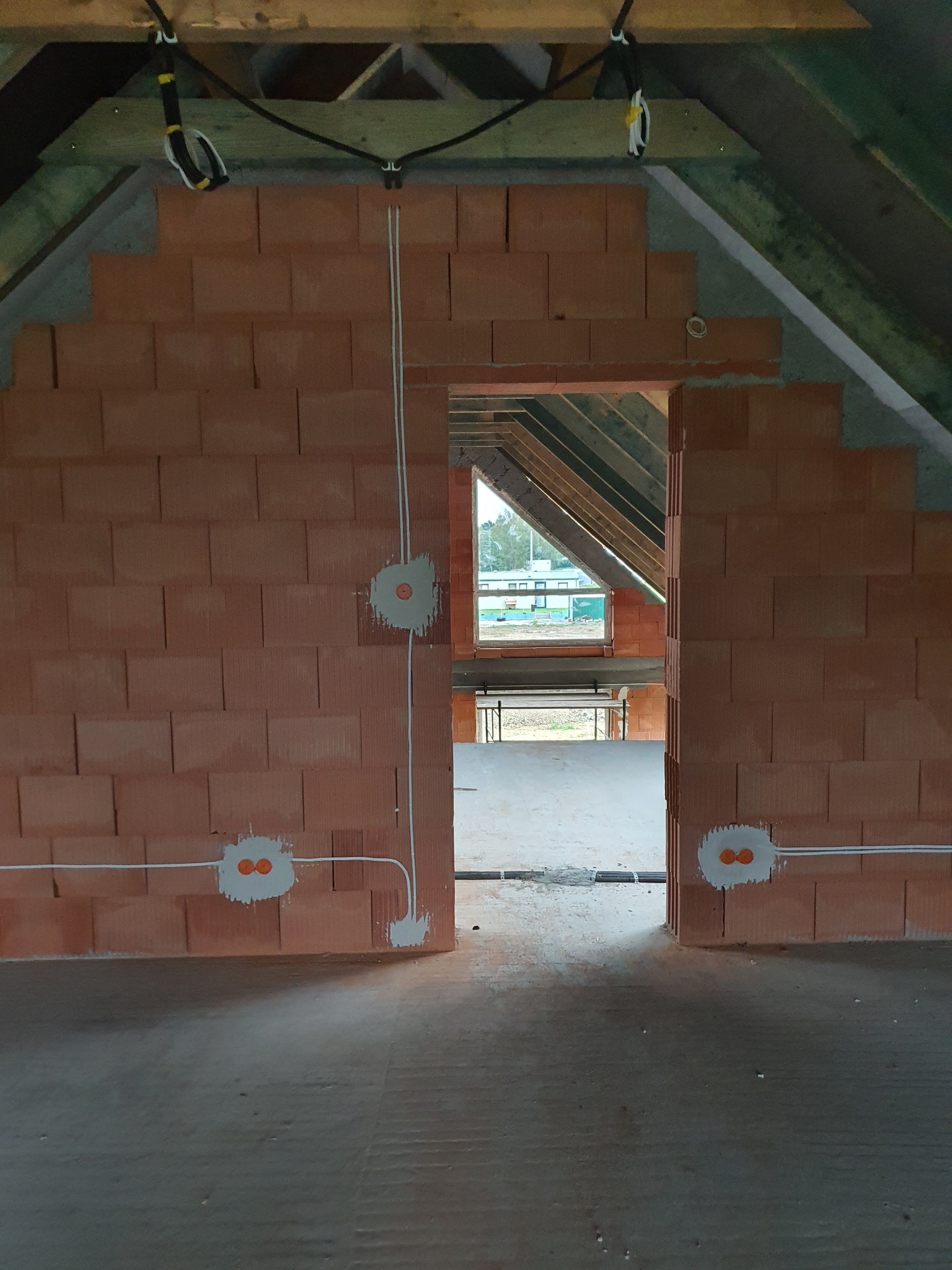 Wnętrze domu w trakcie budowy: ściany z czerwonej cegły, widoczne kable elektryczne w białej osłonie, puszki elektryczne. Surowy stan wykończenia, drewniana konstrukcja dachu.