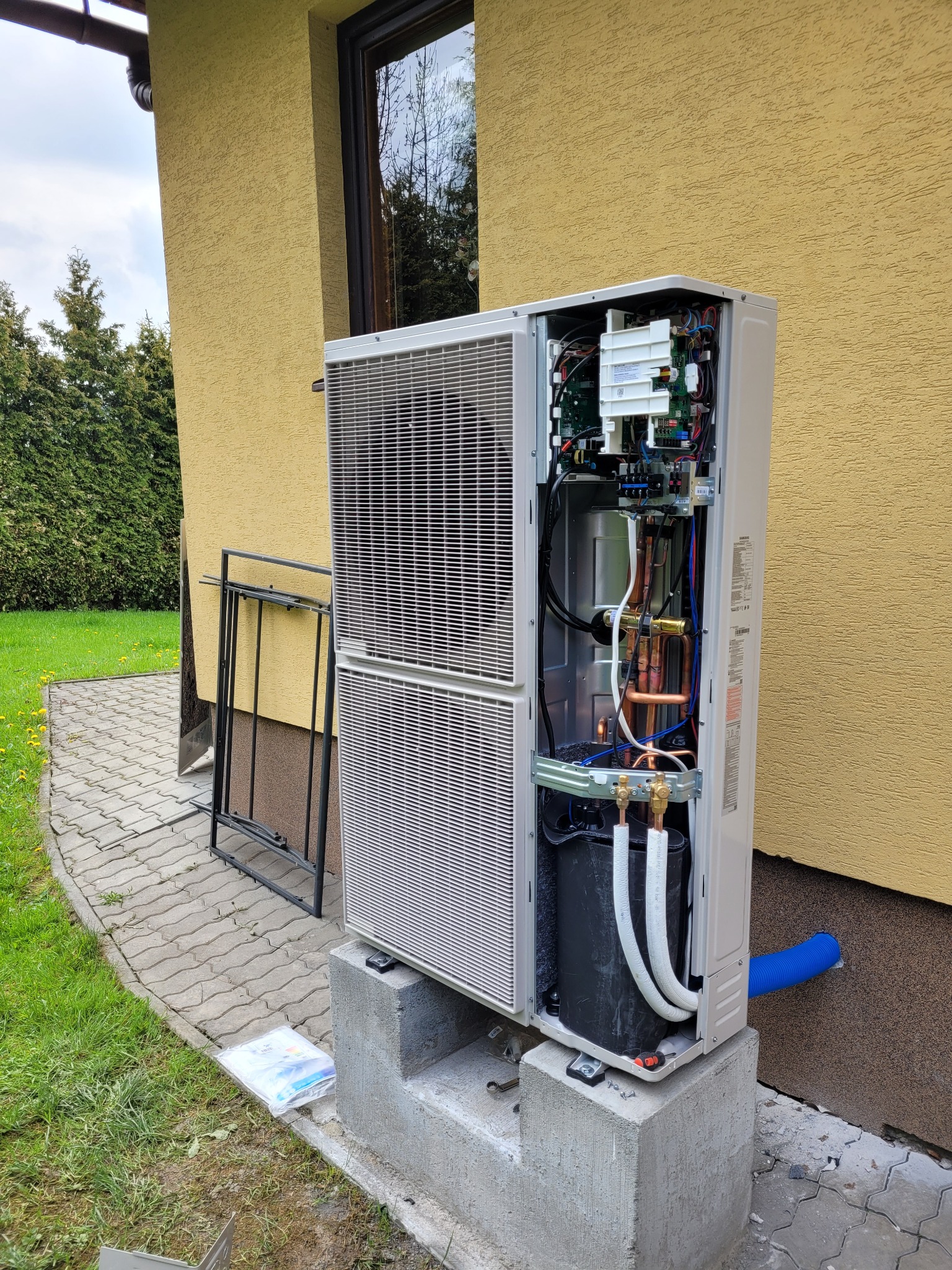 Otwarta pompa ciepła na betonowym fundamencie przy budynku z żółtą elewacją. Widoczne miedziane rury, elektronika i przewody. Obok leżą instrukcje montażu.