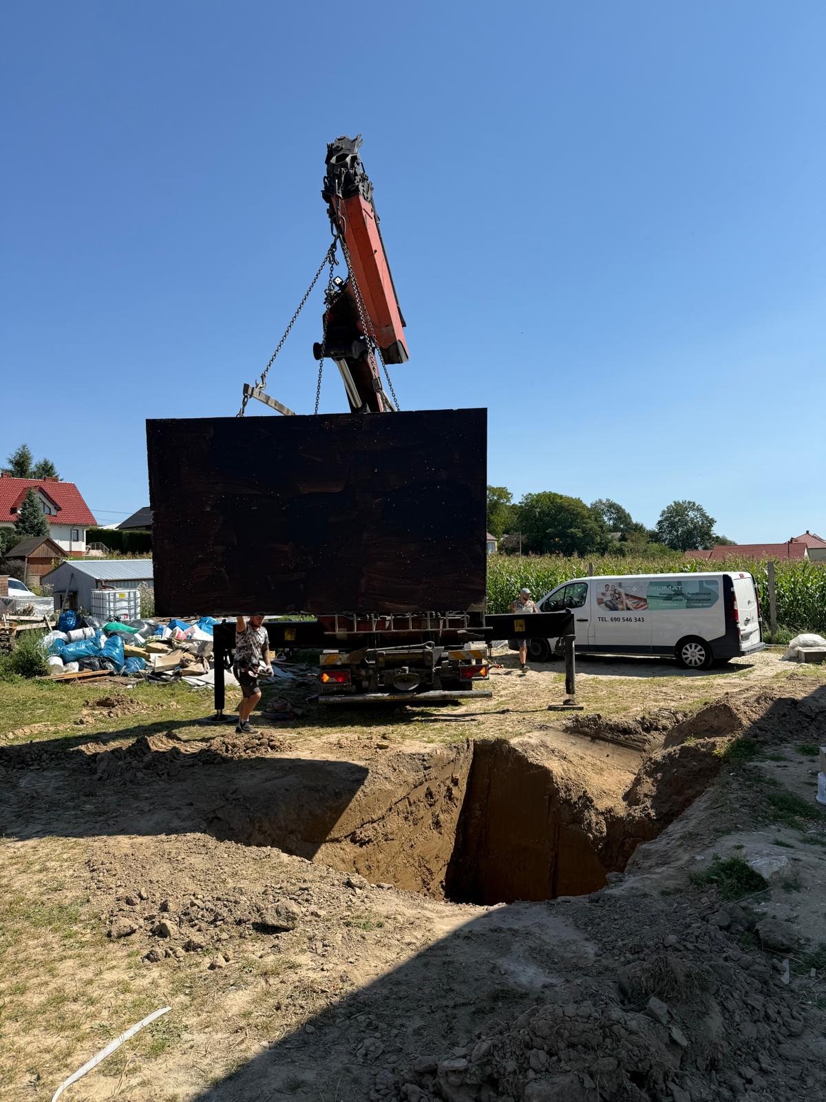 Montaż prefabrykowanej piwnicy za pomocą dźwigu; wykop w ziemi, widoczny samochód dostawczy firmy. Słoneczny dzień, budowa domu.