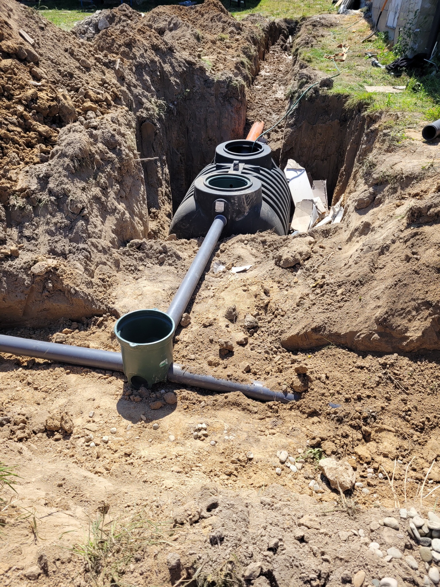 Montaż szamba ekologicznego z rurami w wykopie. Widoczne elementy instalacji i ziemia. Prace w toku, teren budowy.