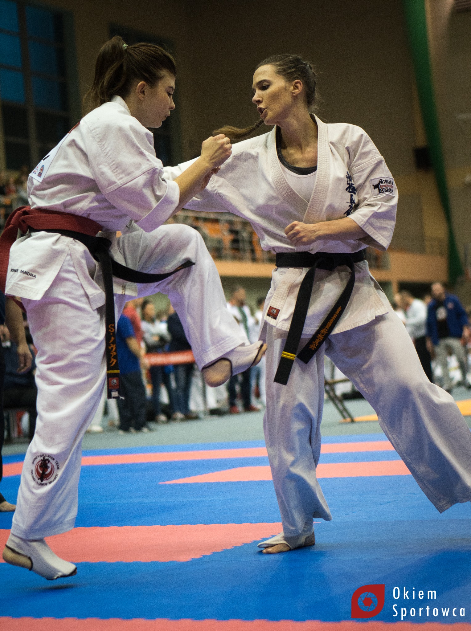 Dwie kobiety w białych kimonach walczą podczas zawodów karate. Jedna z nich wykonuje kopnięcie, druga blokuje. Widoczni widzowie w tle.