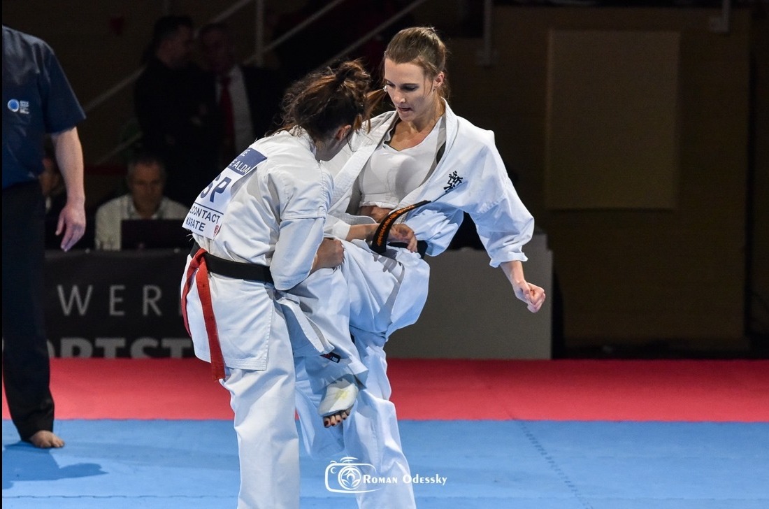 Kobieta w białym kimonie wykonuje kopnięcie podczas zawodów karate, na tle widowni i banerów reklamowych. Dynamiczne ujęcie z akcji.