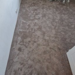 Grzegorz Sadło - Pokój po położeniu brązowej wykładziny dywanowej. Widoczny fragment okna z dekoracjami, grzejnik i fragment żyrandola. Wykładzina o jednolitej strukturze i kolorze.