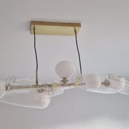Grzegorz Sadło - Nowoczesna, złota lampa sufitowa z trzema białymi kloszami w folii ochronnej, zamontowana na białym suficie. Widoczny fragment ściany i obrazek w ramce.