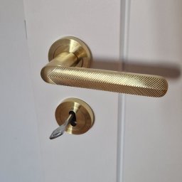 Grzegorz Sadło - Białe drzwi z zamontowaną złotą klamką o teksturowanej powierzchni i kluczem w zamku, ukazujące dbałość o detale wykończenia wnętrza.